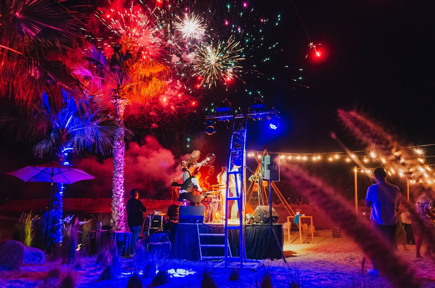 Colorful fireworks display illuminating palm trees and stage at night beach party.