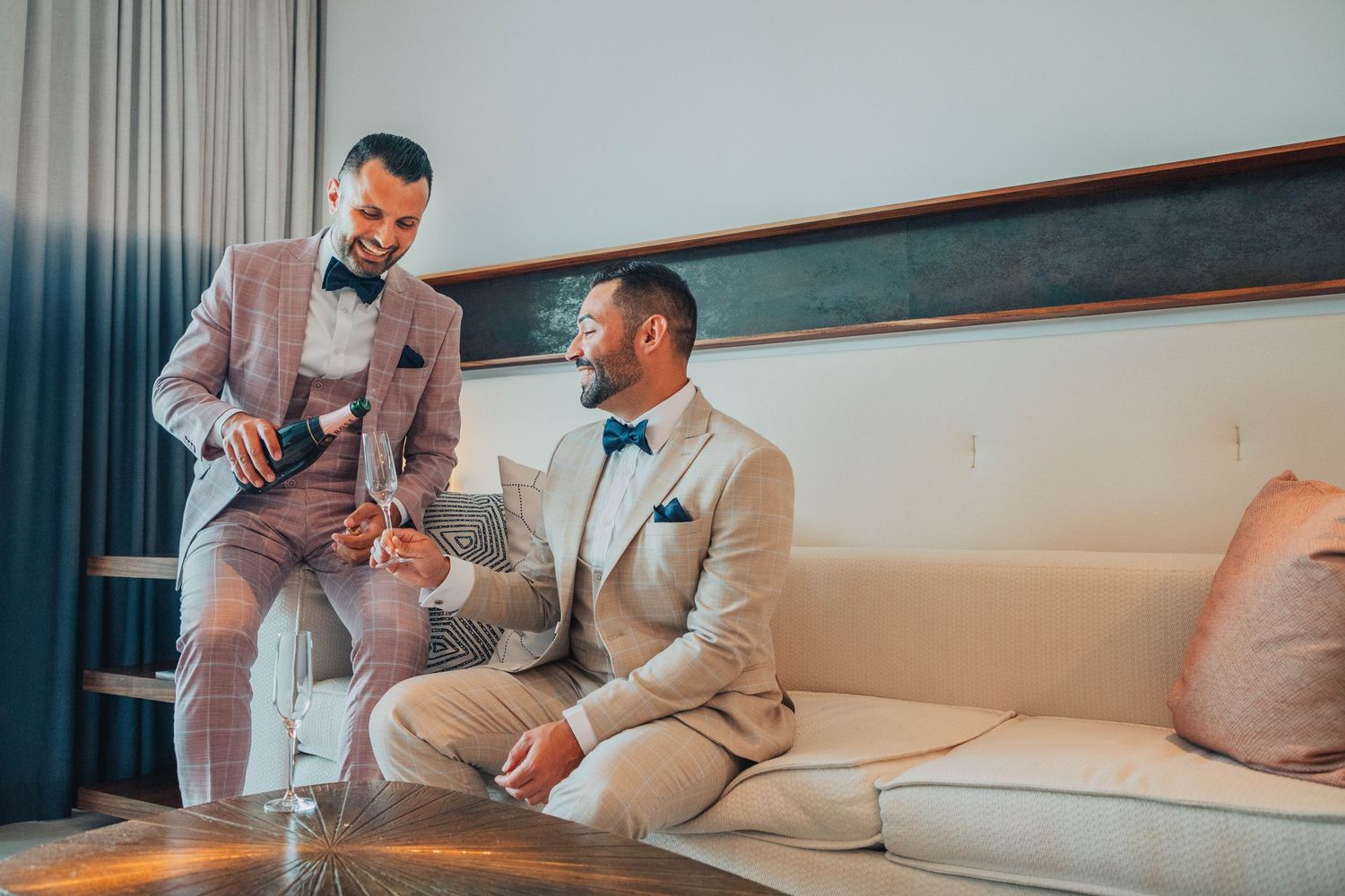Two elegantly dressed people in light colored suits conversing in a modern living room setting.