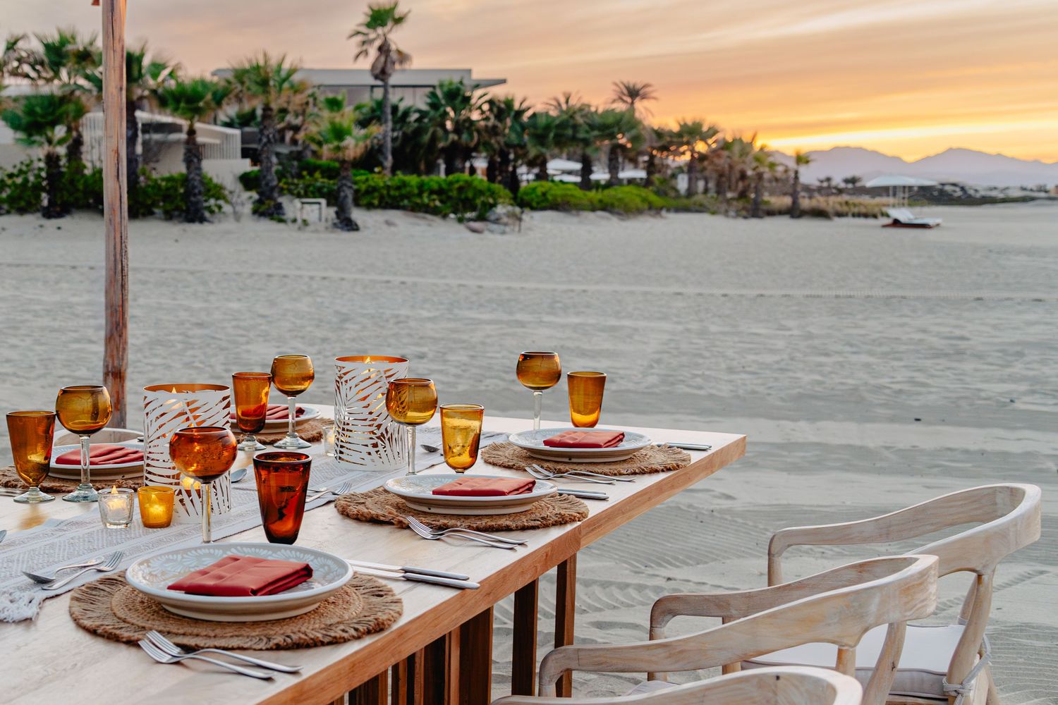 Outdoor dining tables set with glassware and place settings overlooking a waterfront at sunset.