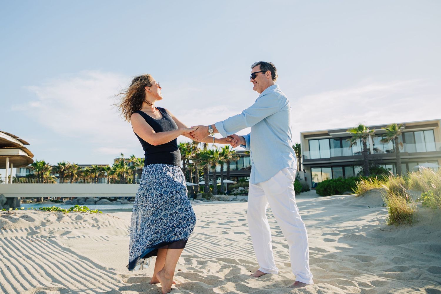 A couple dances together on a beach boardwalk with coastal homes in the background on a sunny day.