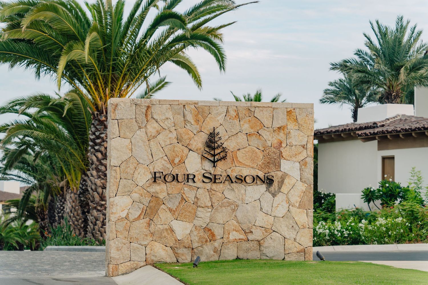 Tropical landscaping with palm trees and stone walls line a luxury resort entrance with Four Seasons signage.