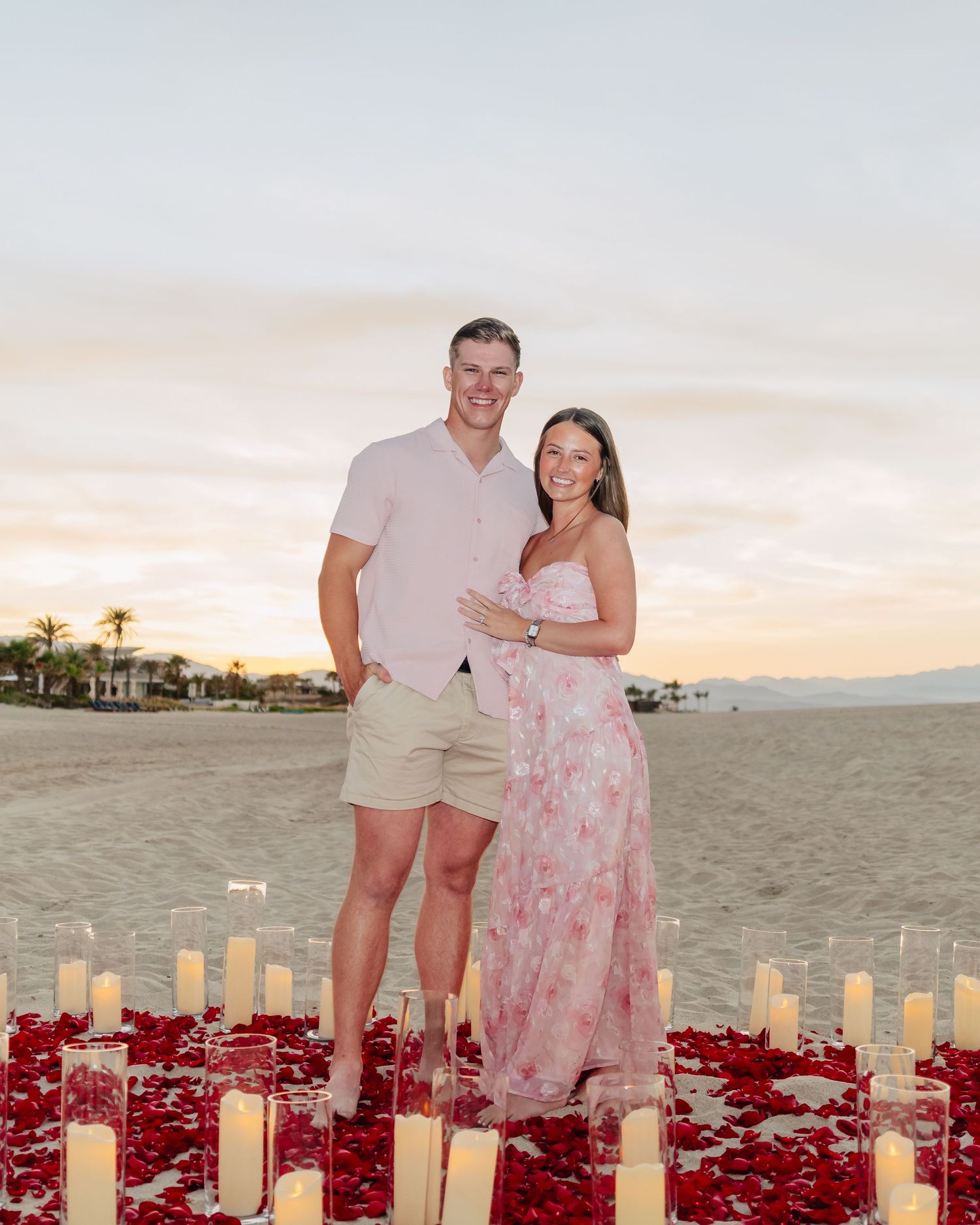 Couple standing together on beach with romantic candle and rose petal setup at sunset.