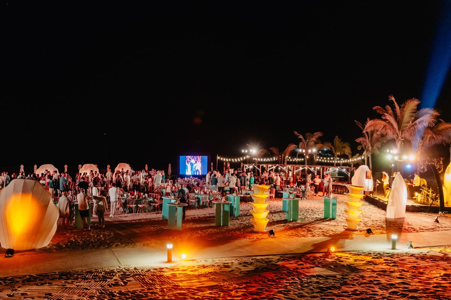 Beachfront evening celebration with illuminated palm trees and string lights creating a festive atmosphere.