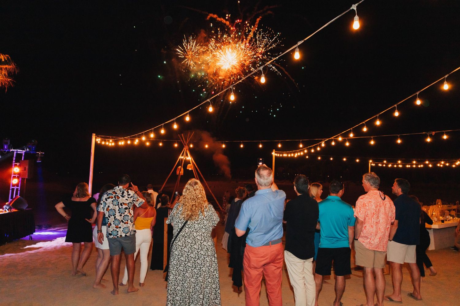 Crowd gathered under string lights watching fireworks display at beach party.