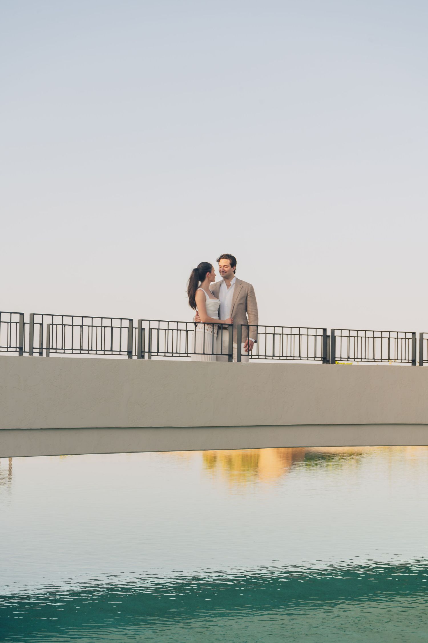 A couple shares an intimate moment while standing on a bridge over turquoise waters at sunset.