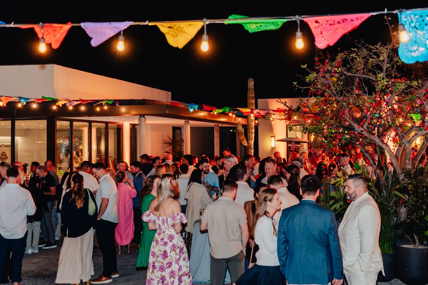 Outdoor evening event with string lights and colorful bunting decorations hanging above a crowd of party guests.