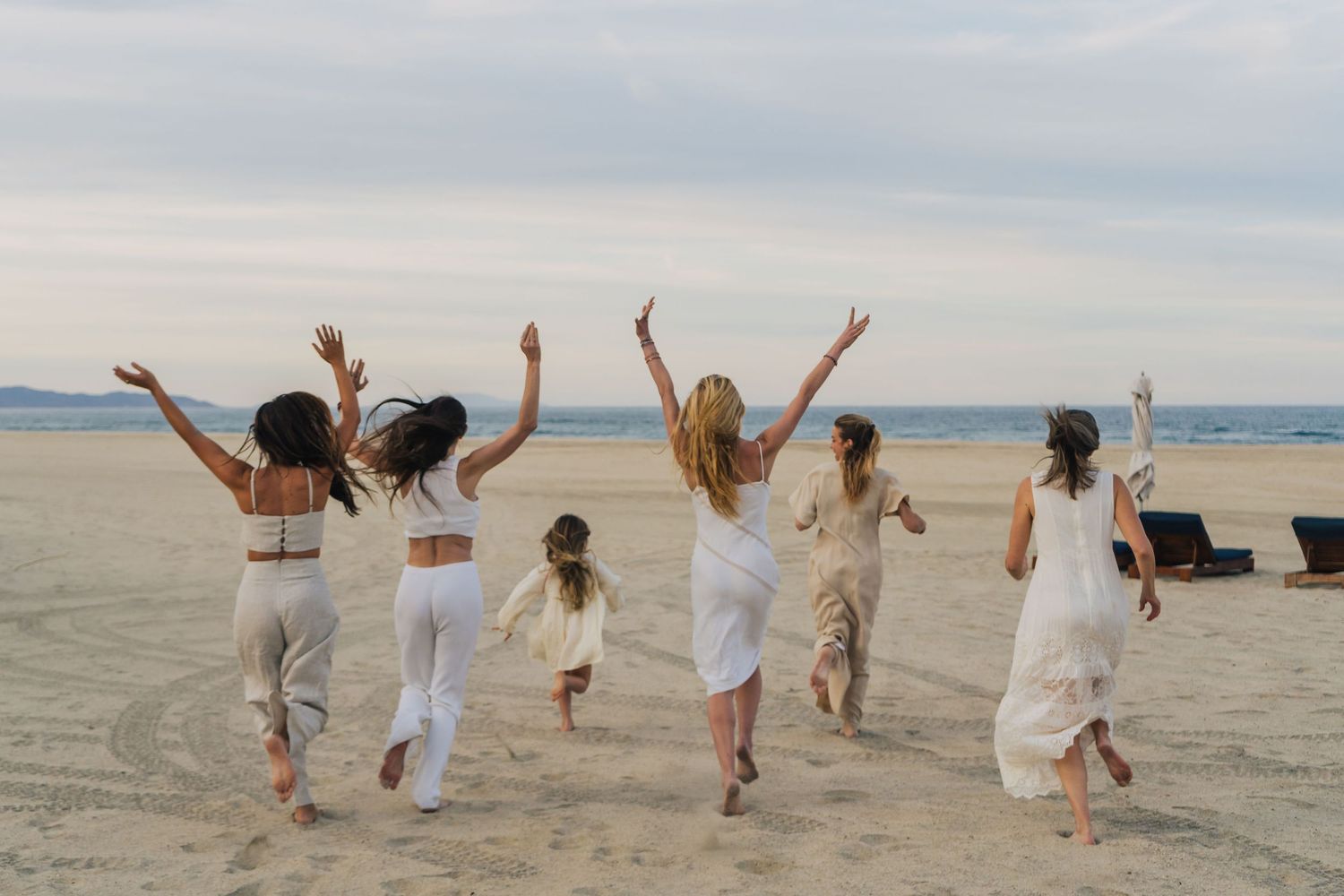 A group of people in white outfits joyfully jumping on a beach at sunset with mountains in the background.