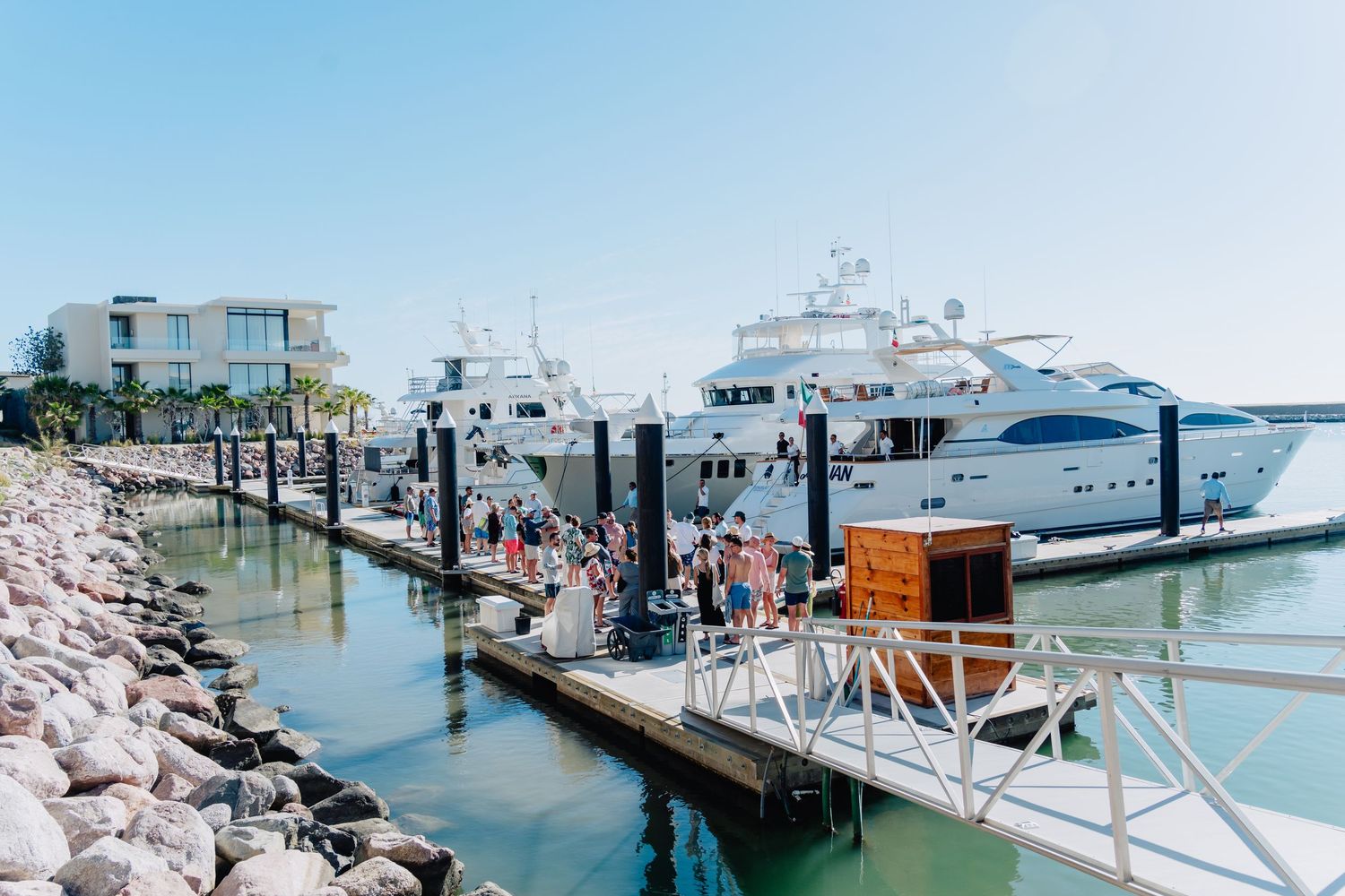 Luxury yachts docked at a marina with people walking along the wooden pier on a sunny day.