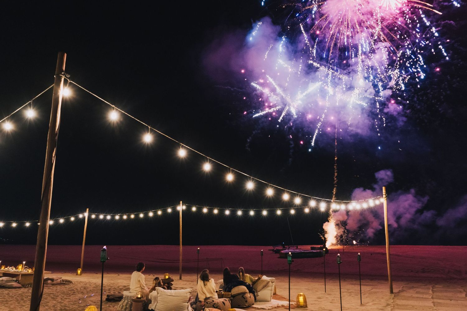 Colorful fireworks illuminate the night sky over a beach celebration with string lights creating a magical atmosphere.