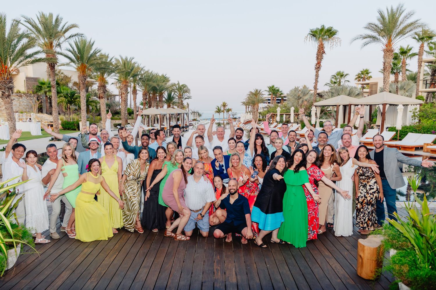 Large group of wedding guests gather for a panoramic photo at a tropical resort with palm trees and greenery.