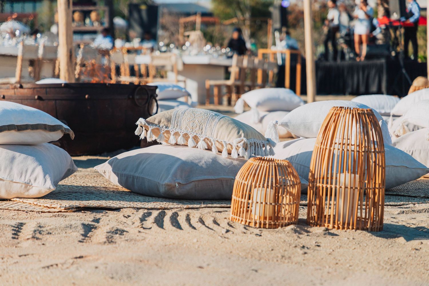 Beach lounge setup with wicker lanterns and white cushions on sandy shore.