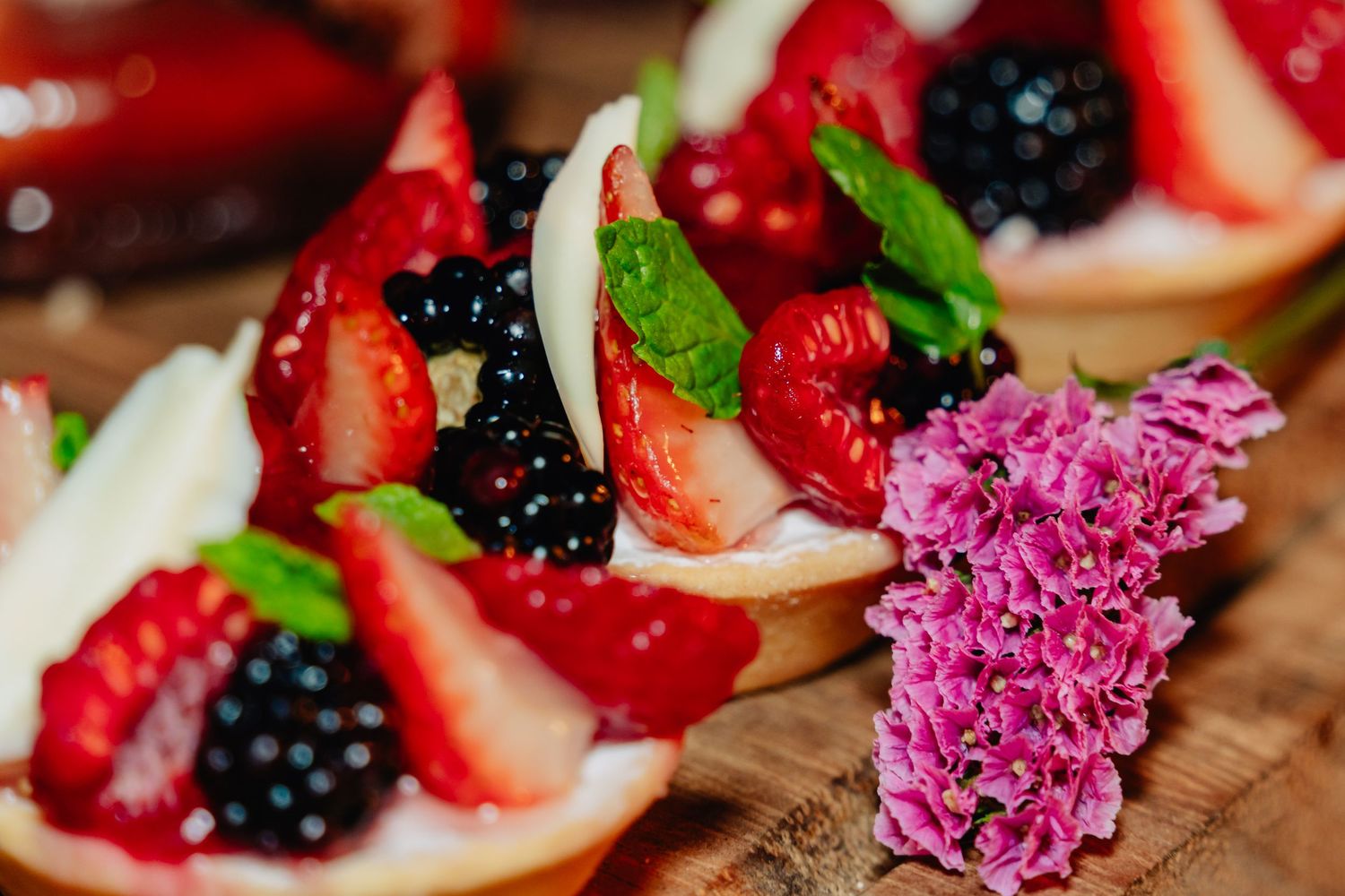 Fresh berry tarts with strawberries and blackberries garnished with mint leaves and purple lilac on wooden surface.