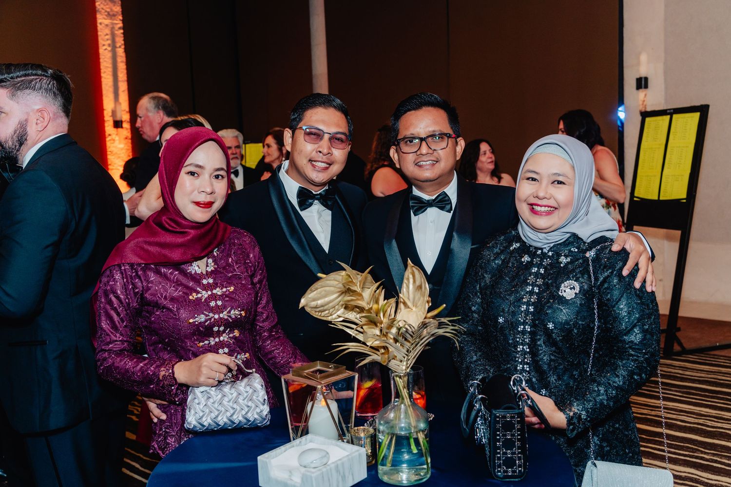 A group of formally dressed guests at an elegant event pose together in front of a decorated venue.