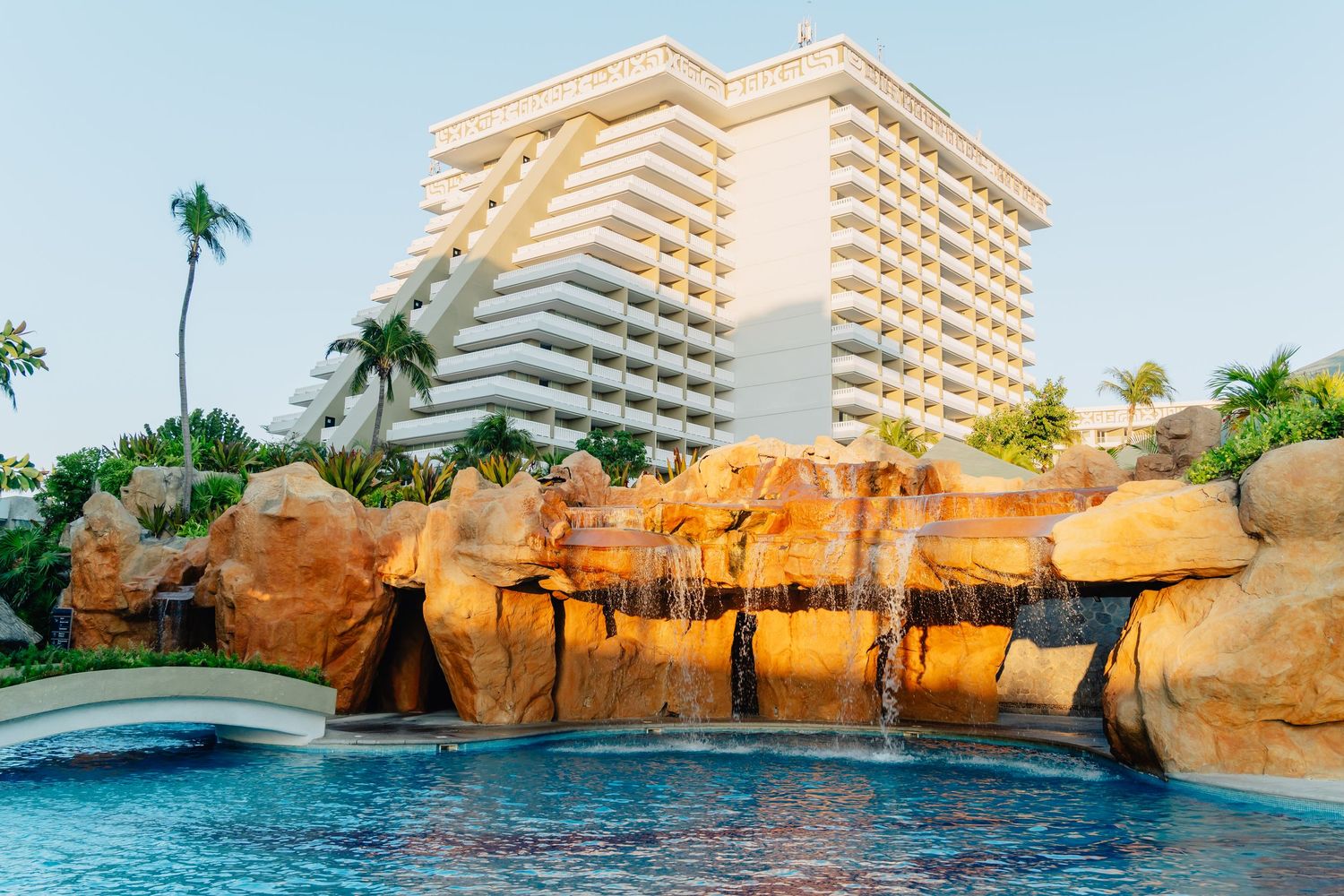 Resort swimming pool with dramatic rock waterfall feature against modern hotel architecture.