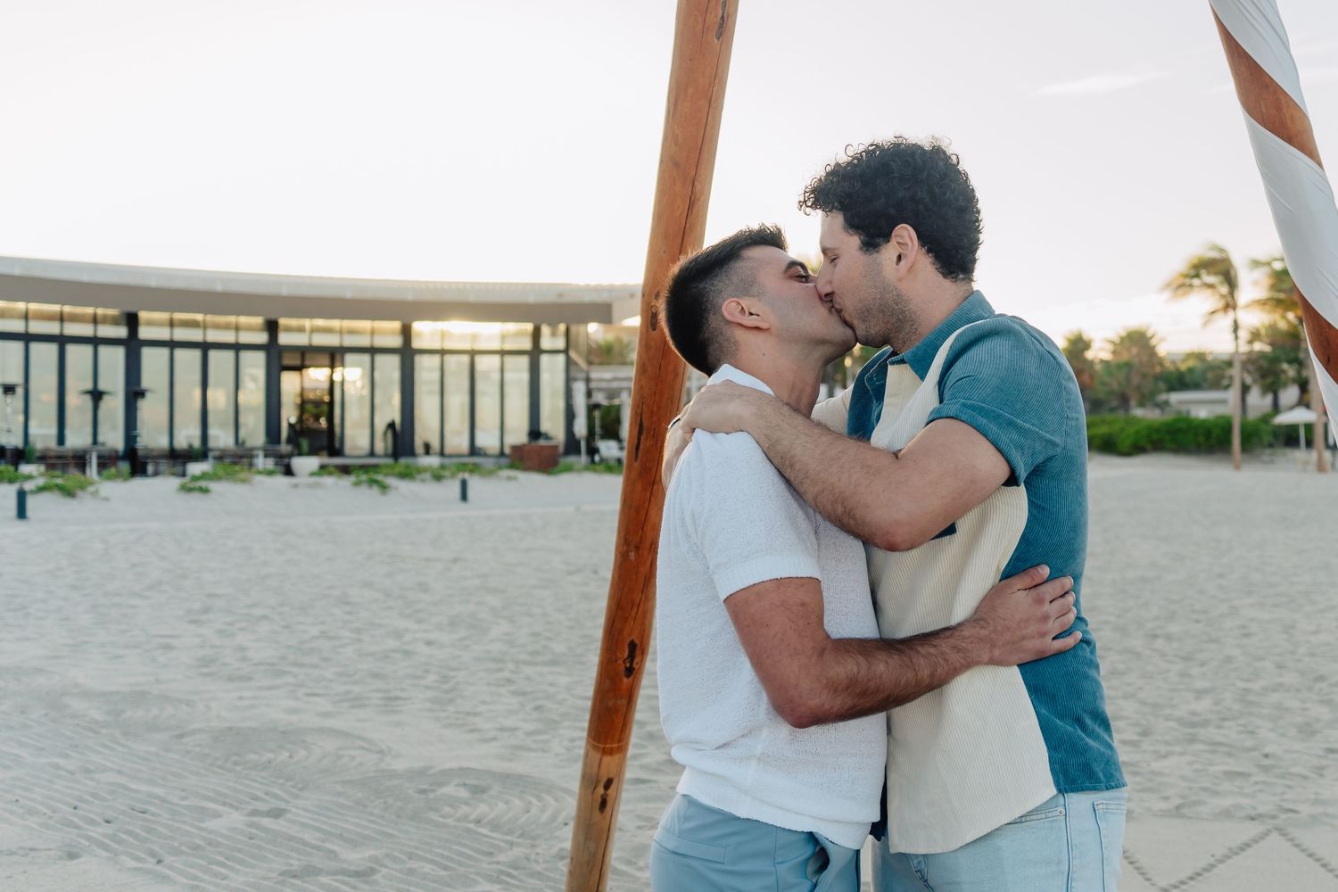 Couple sharing intimate moment under wooden beach teepee at golden hour.