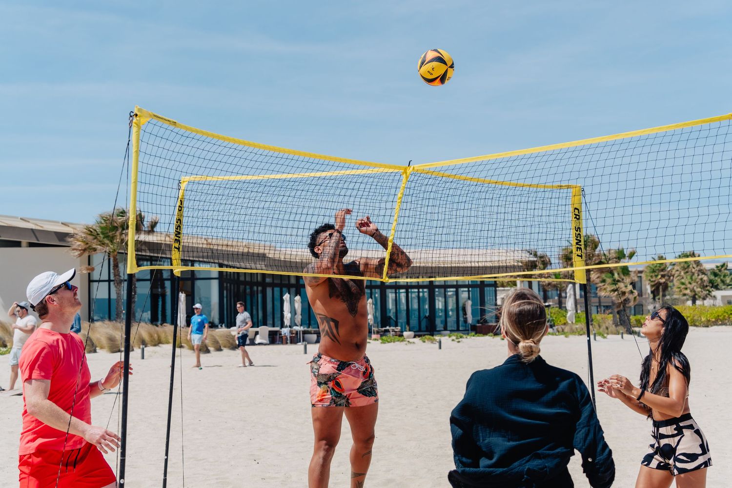 Beach volleyball game in progress with players jumping to hit ball over net.