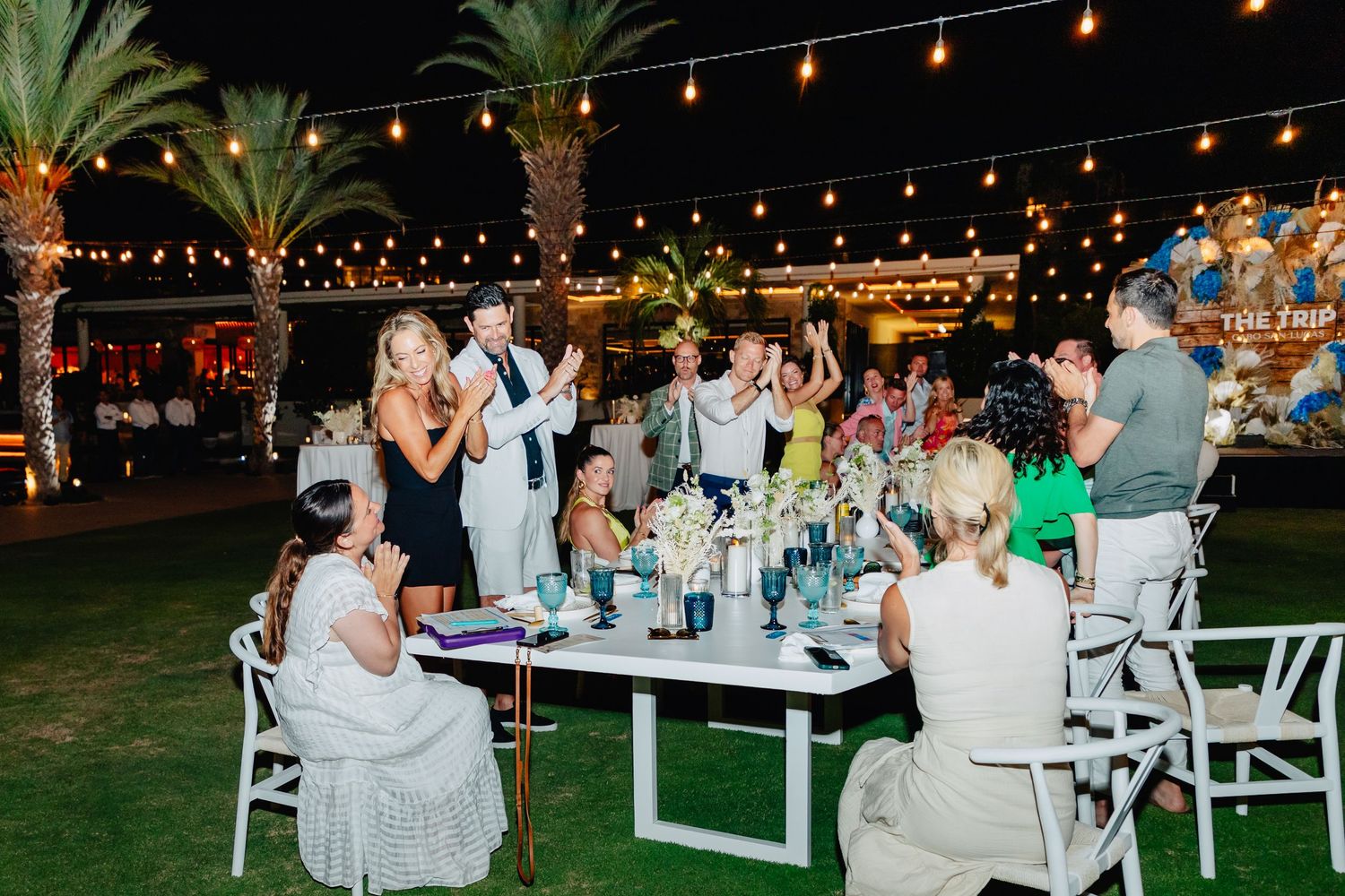 Guests enjoy an outdoor evening reception with string lights and palm trees at an elegant dinner party.