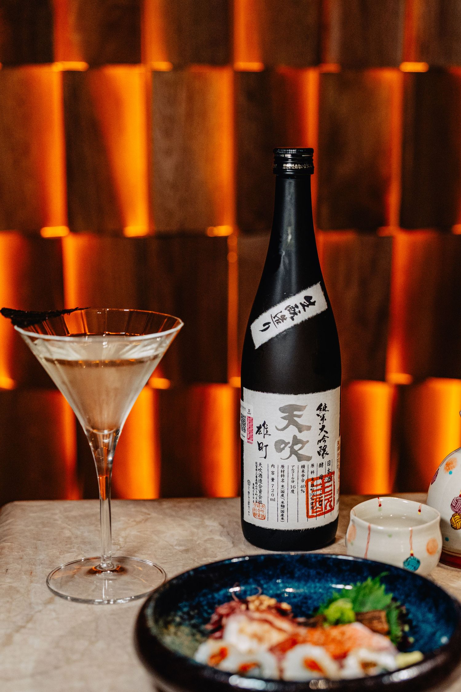 Elegant bar setting with Japanese sake bottles and martini glasses on marble counter with orange ambient lighting.