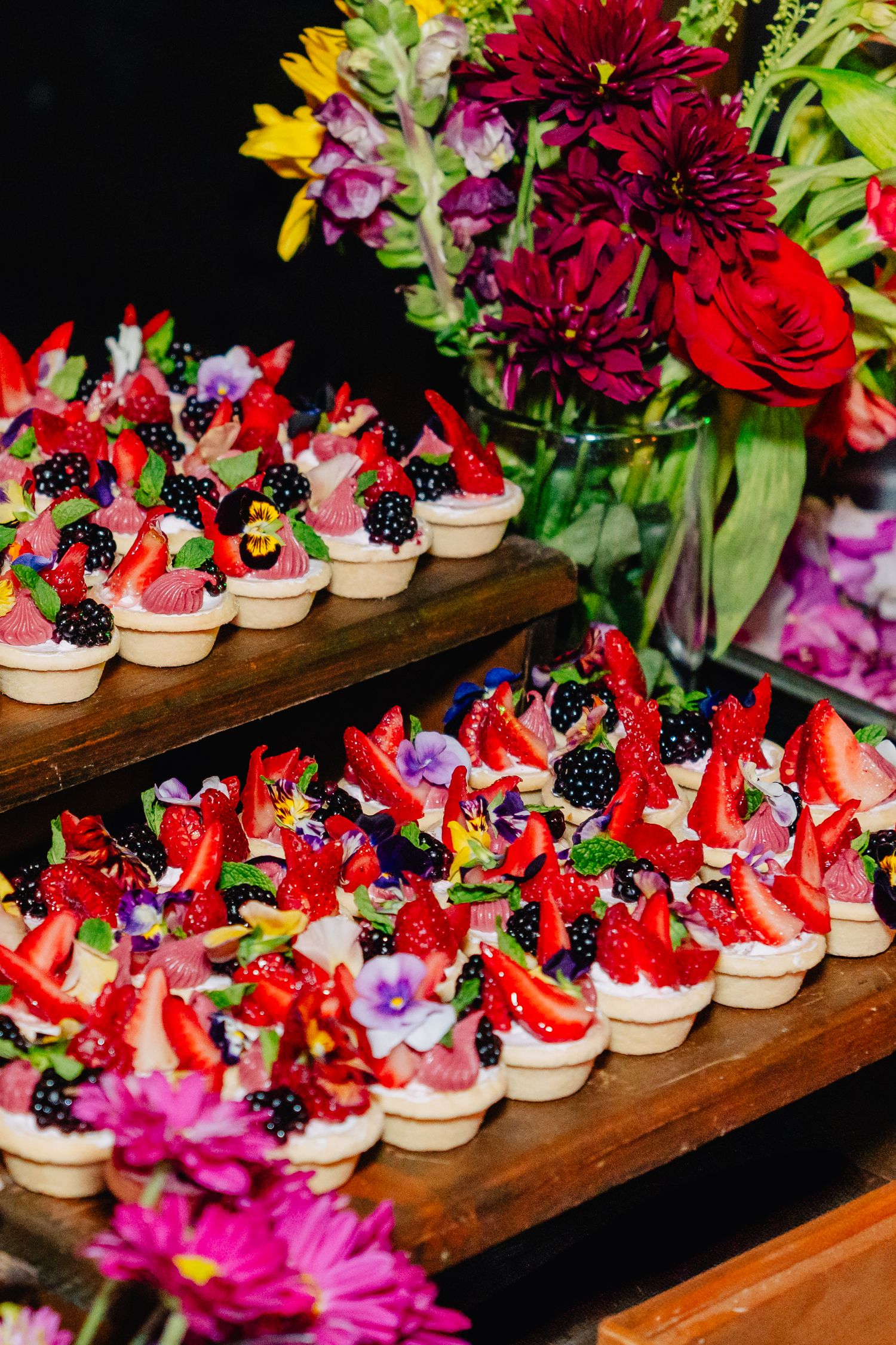 Rows of colorful mini desserts decorated with fresh berries and flowers displayed on wooden shelves.