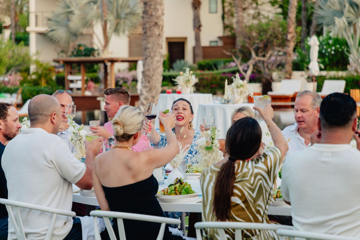 Wedding guests raise their glasses in a joyful toast at an outdoor reception with palm trees in the background.