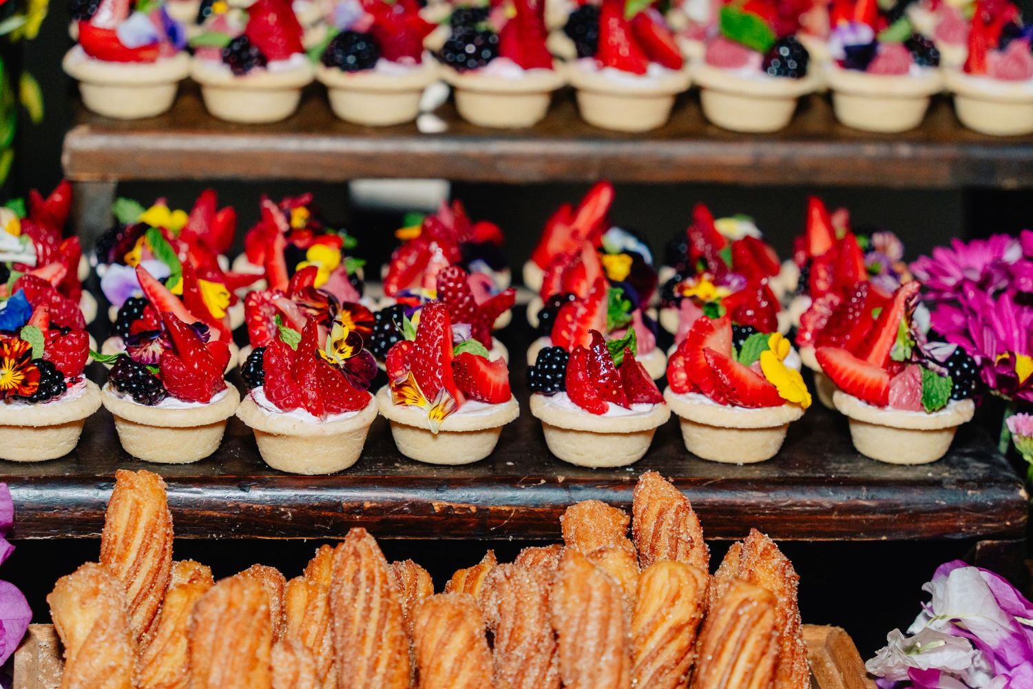 Rows of elegant desserts including chocolate tarts with fresh berries and golden pastries arranged on dark display trays.