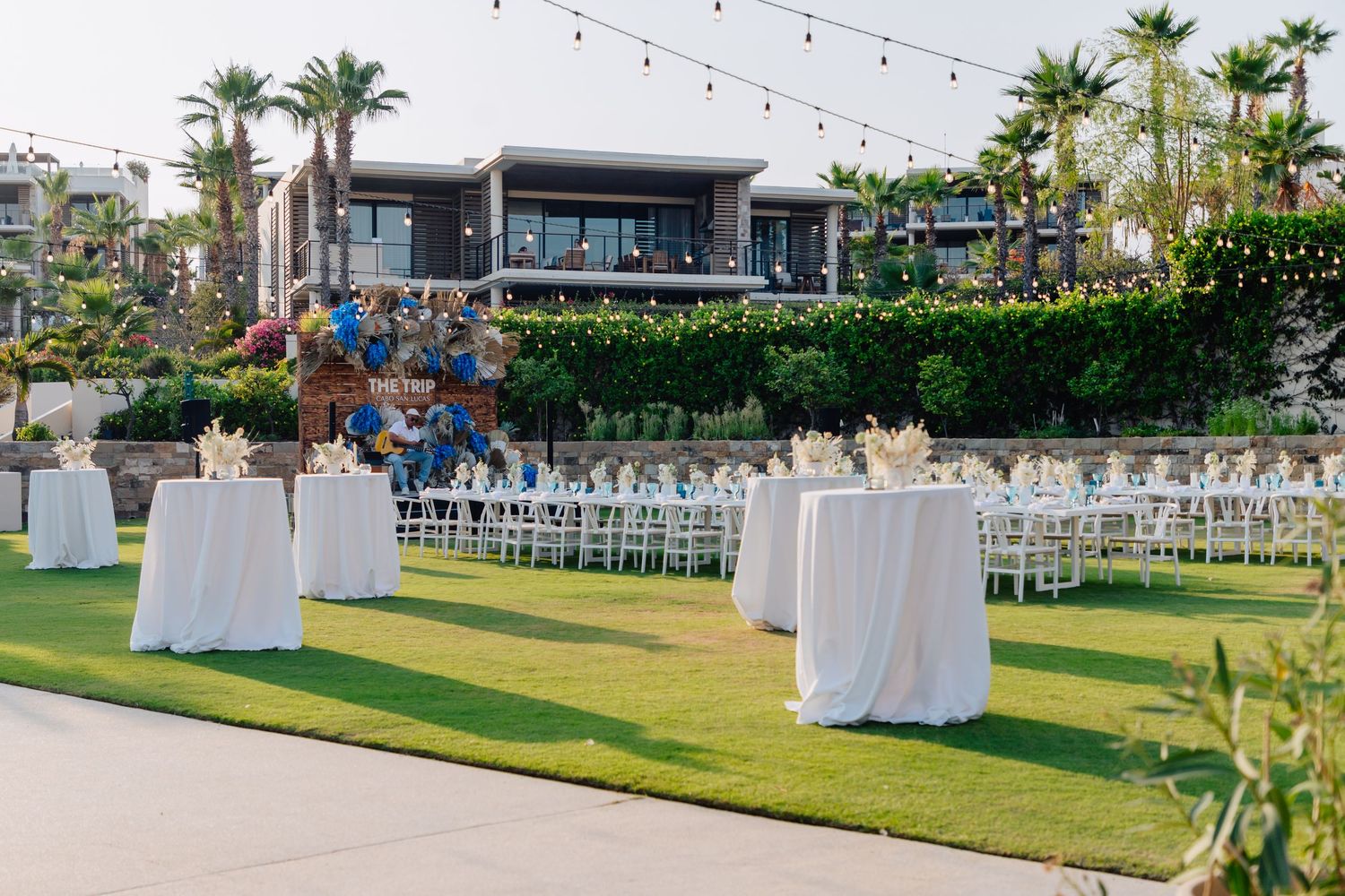 Long white reception tables arranged on a manicured lawn with palm trees in the background.