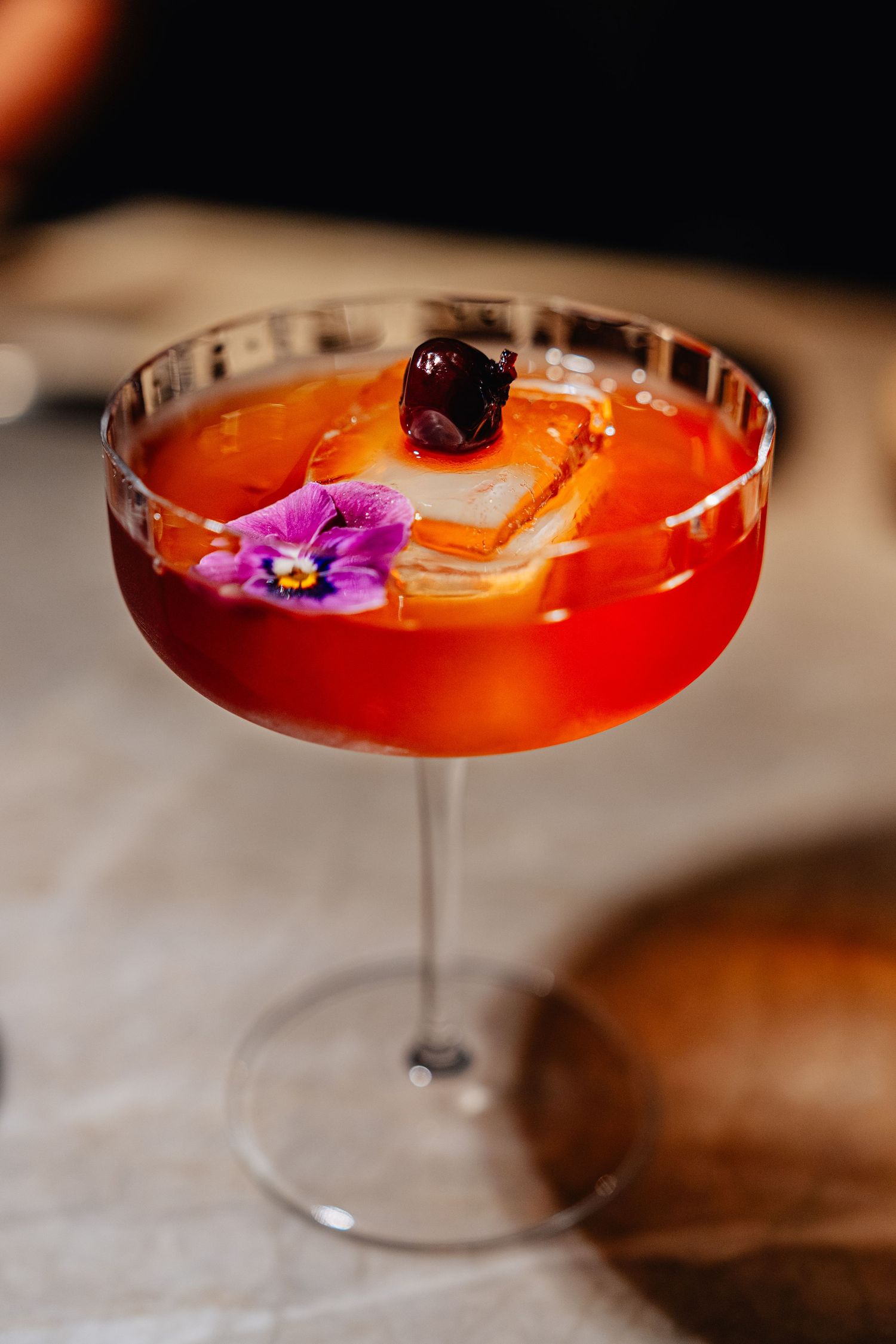 Vibrant craft cocktail garnished with edible flowers and cherries in a coupe glass against a dark background.