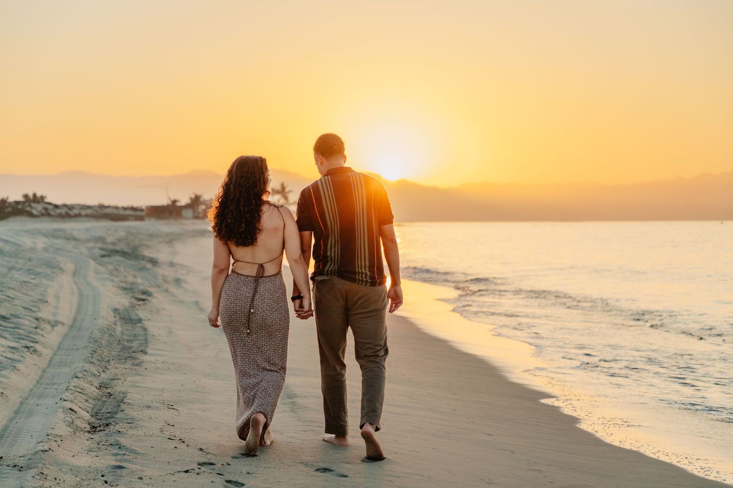 Couple walking hand in hand on a sandy beach at sunset with golden sunlight reflecting off the ocean waves.