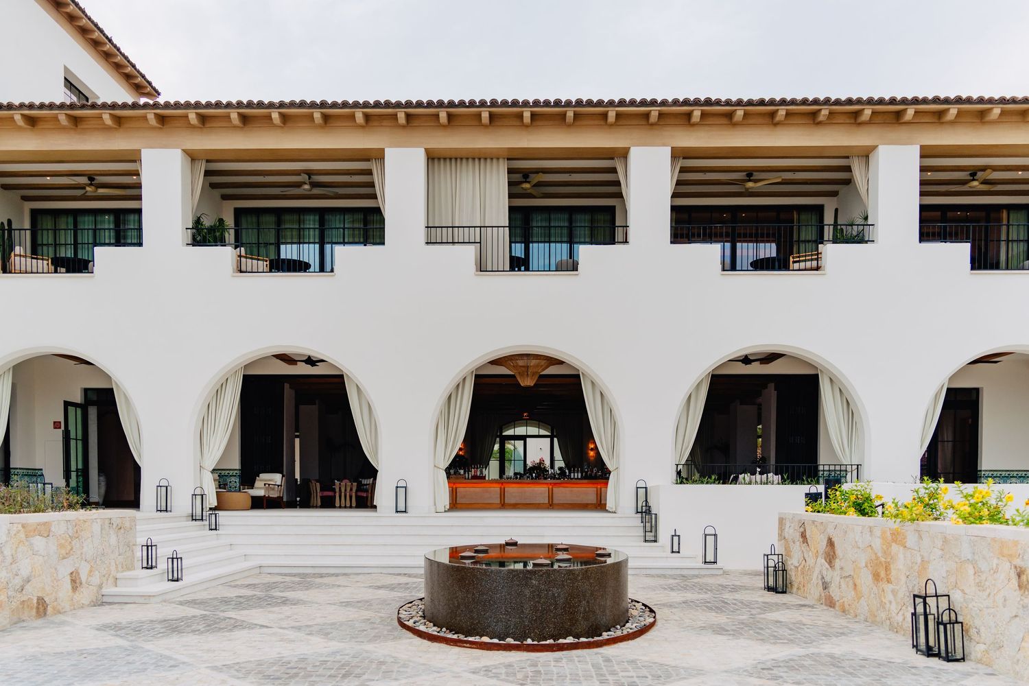 Mediterranean-style resort entrance featuring white arched doorways and a circular stone fountain centerpiece.