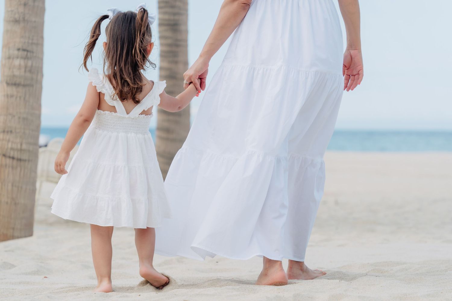 People in white dresses walk along a tropical beach with palm trees and ocean views.