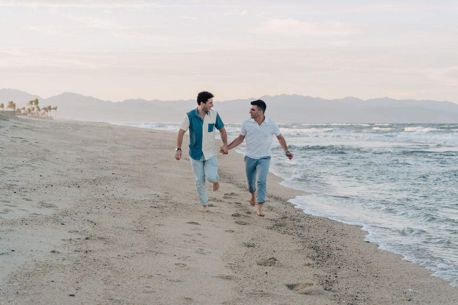 A couple walks hand in hand along a sandy beach with mountains and ocean waves in the background at sunset.