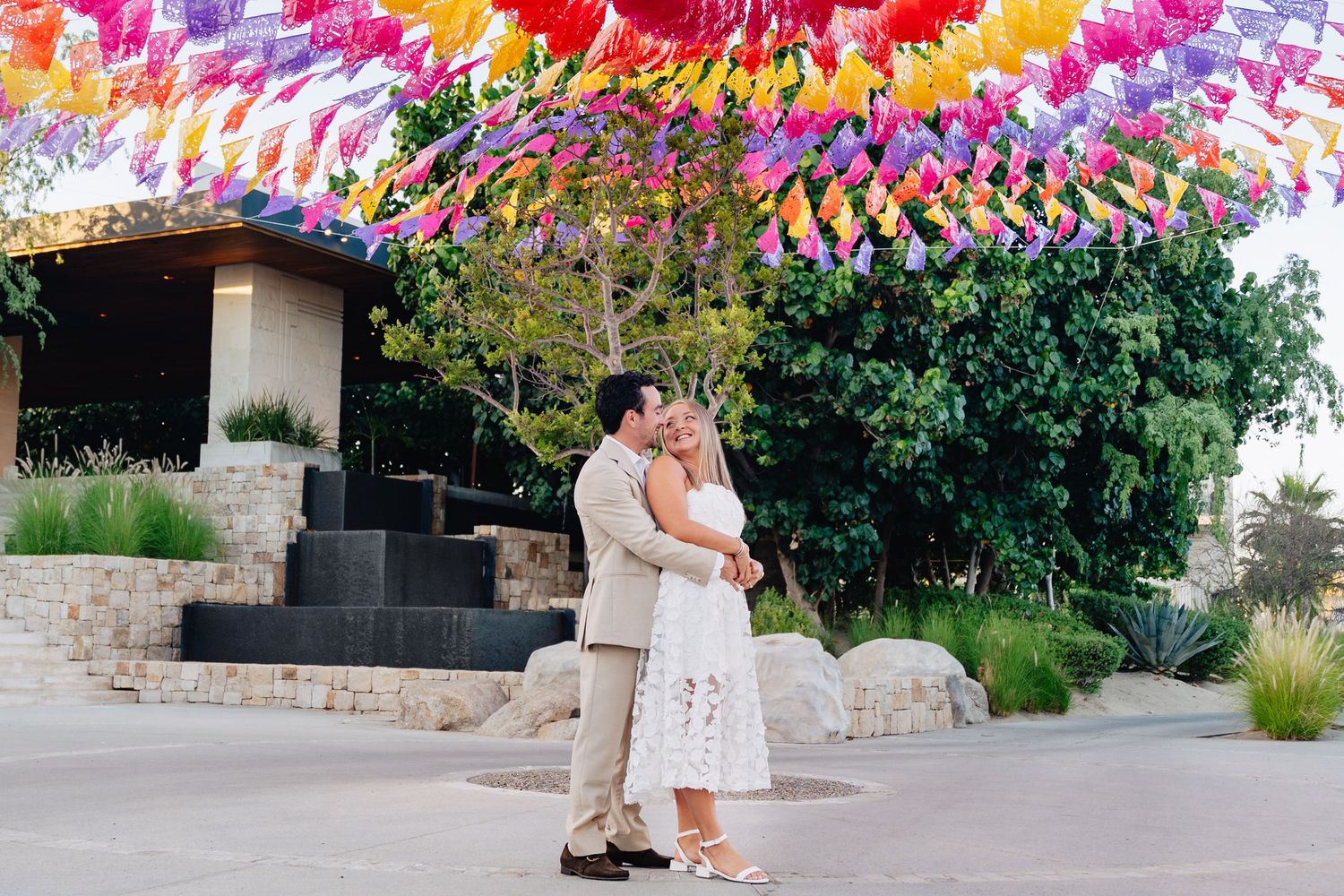 A couple shares a romantic moment under vibrant pink and orange floral decorations at an outdoor venue.
