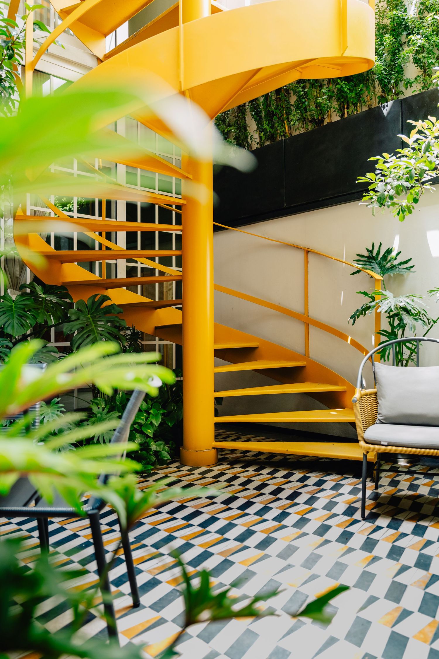 Yellow spiral staircase with geometric patterned floor tiles surrounded by green indoor plants in a bright interior space.