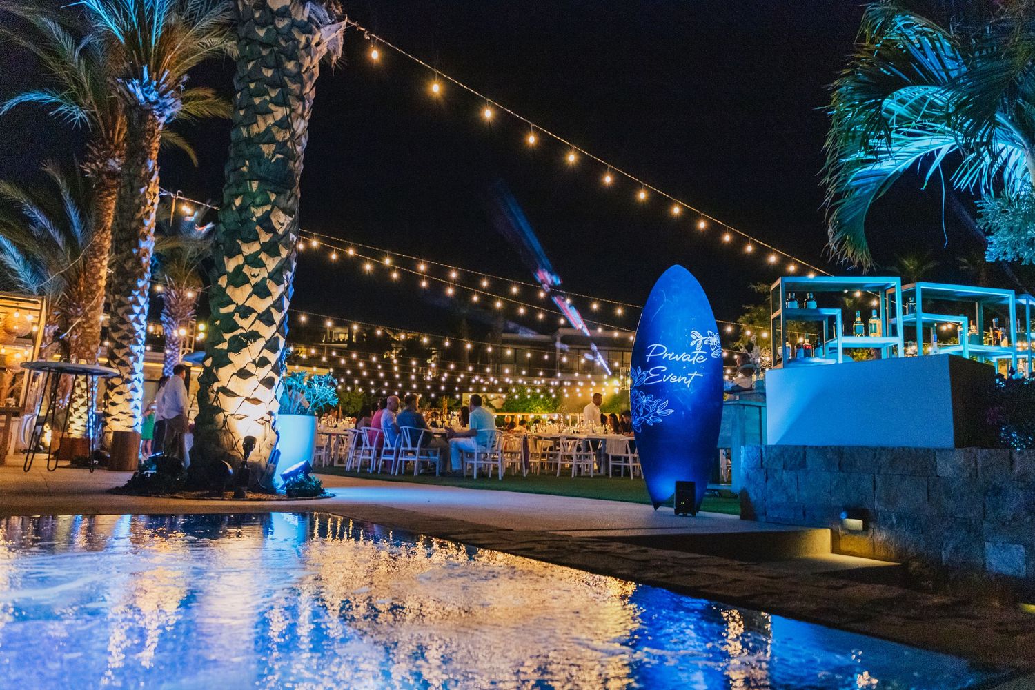 Evening poolside scene with illuminated palm trees and string lights creating romantic atmosphere.