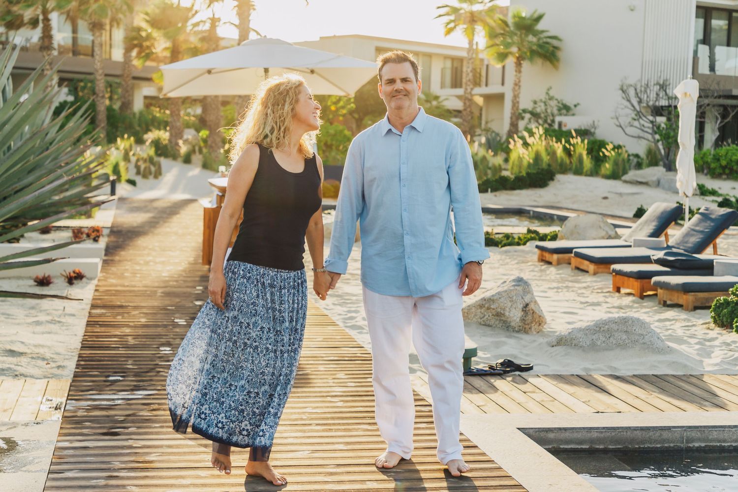 Couple walking hand in hand on wooden boardwalk at tropical resort during sunset.