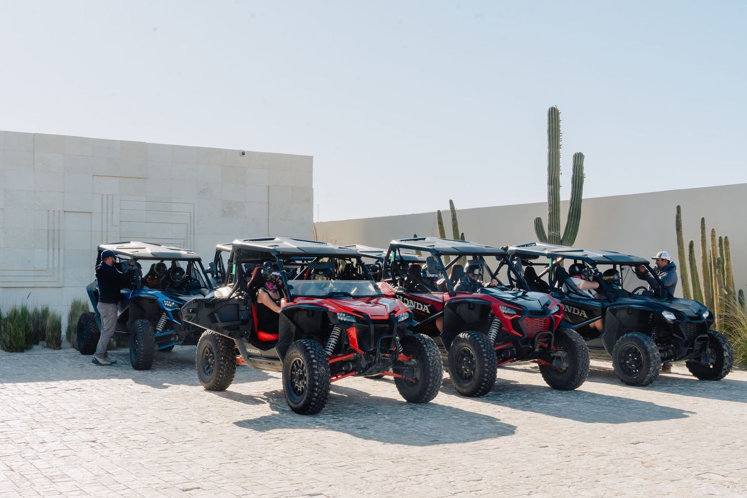 A line of colorful off-road UTVs parked in front of a desert building with a cactus in the background.