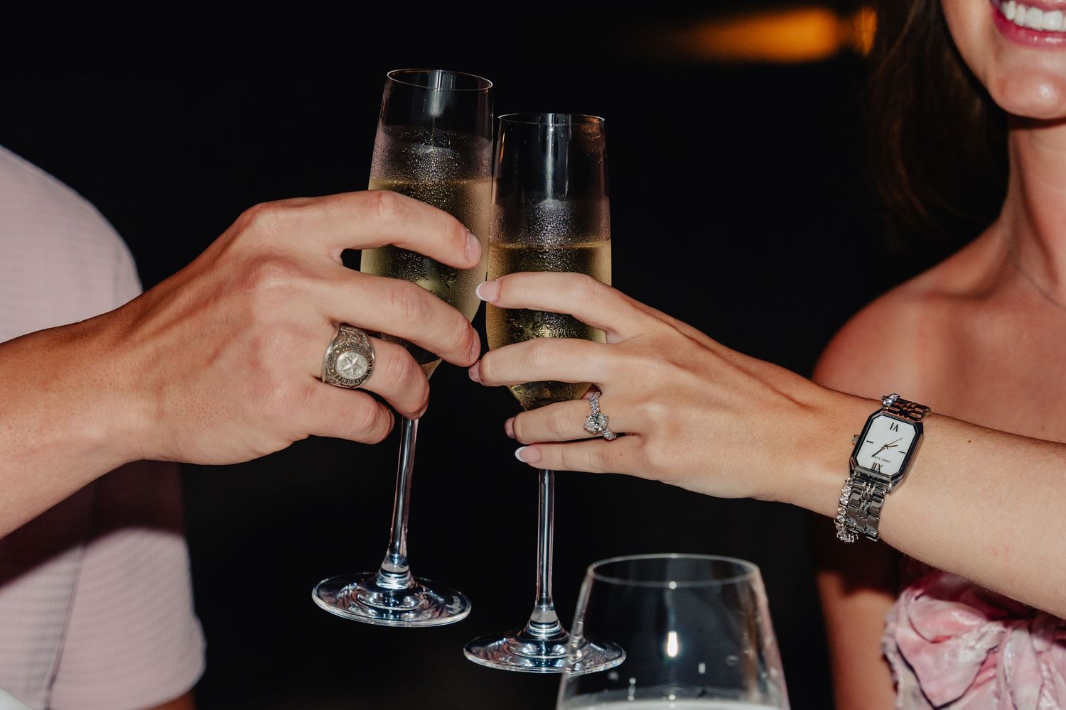 Two hands holding champagne glasses with engagement rings visible.