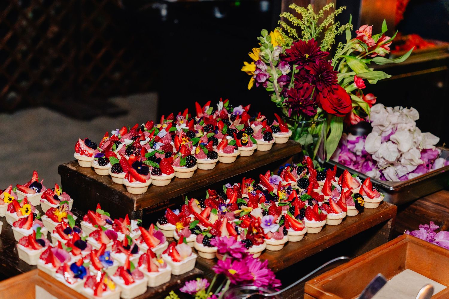 Wooden board displaying rows of colorful decorated desserts garnished with berries and flowers.