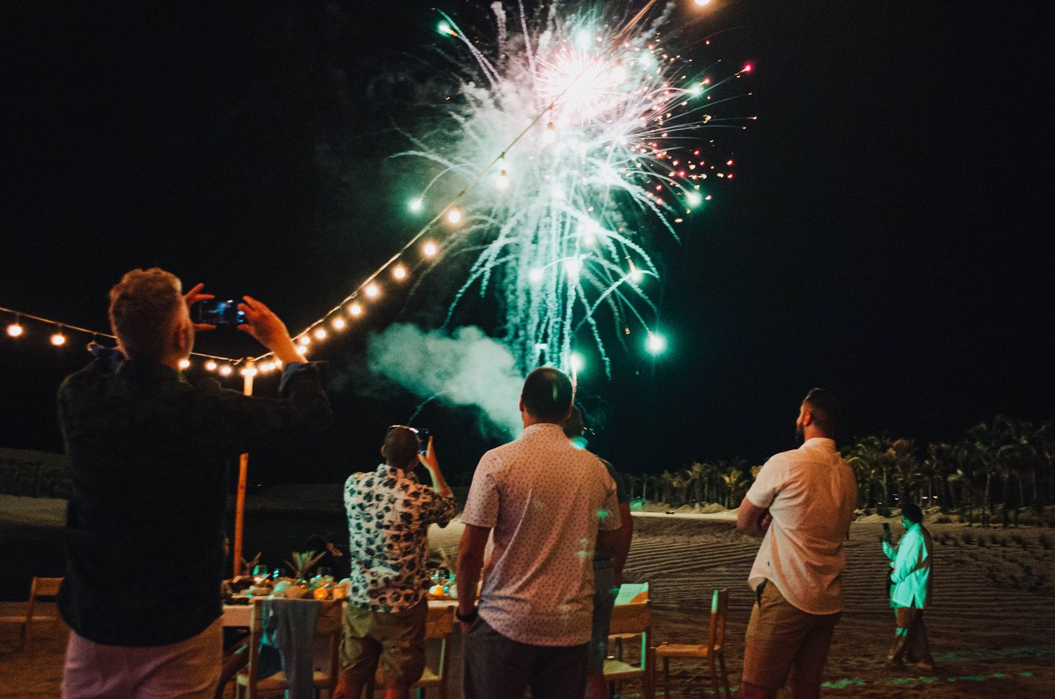 Spectators watching bright fireworks display against night sky with string lights.
