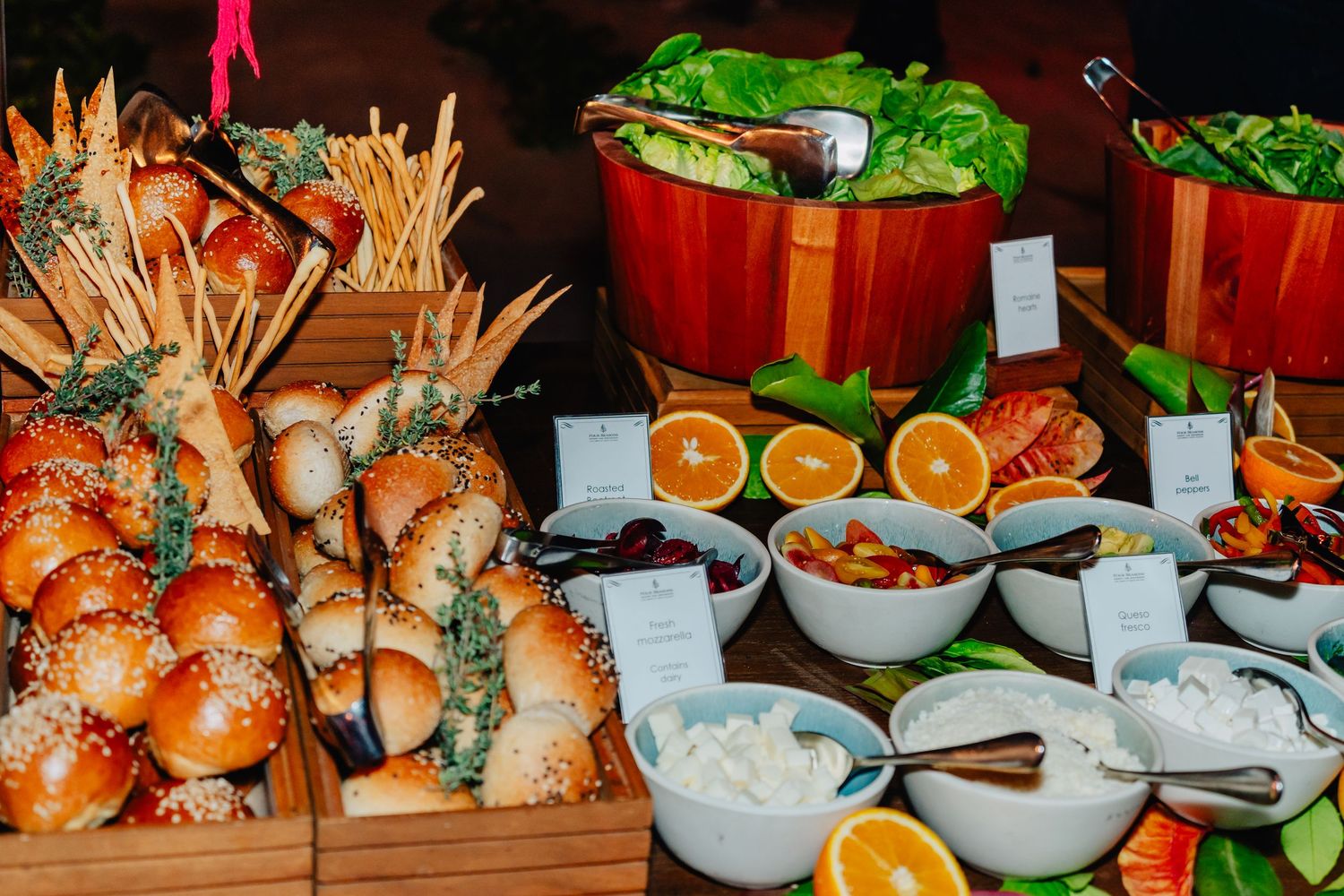 A catering buffet display featuring sliders, salads, citrus fruits and various dipping sauces arranged on a wooden table.