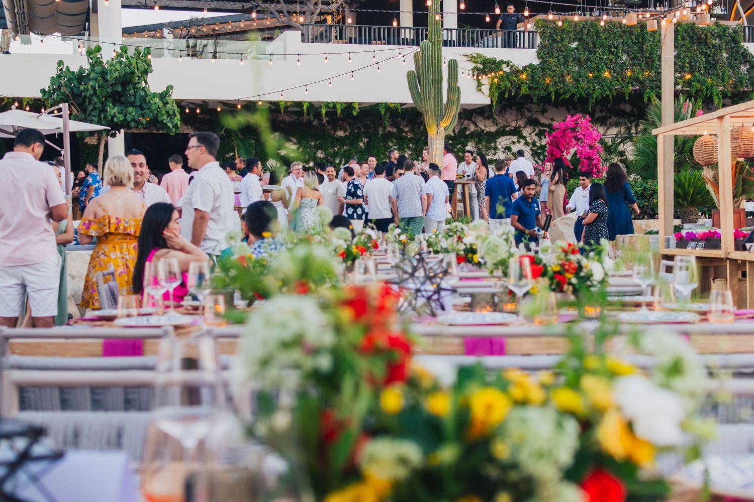Outdoor daytime event setup with colorful floral centerpieces and table settings under palm trees.