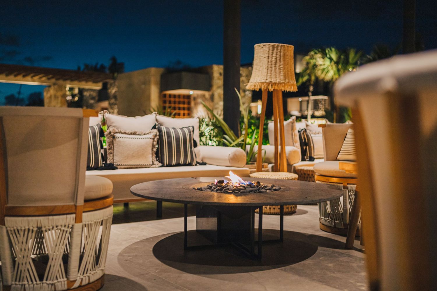 Cozy outdoor patio seating area with woven chairs and a round fire pit table illuminated by wooden table lamps at dusk.