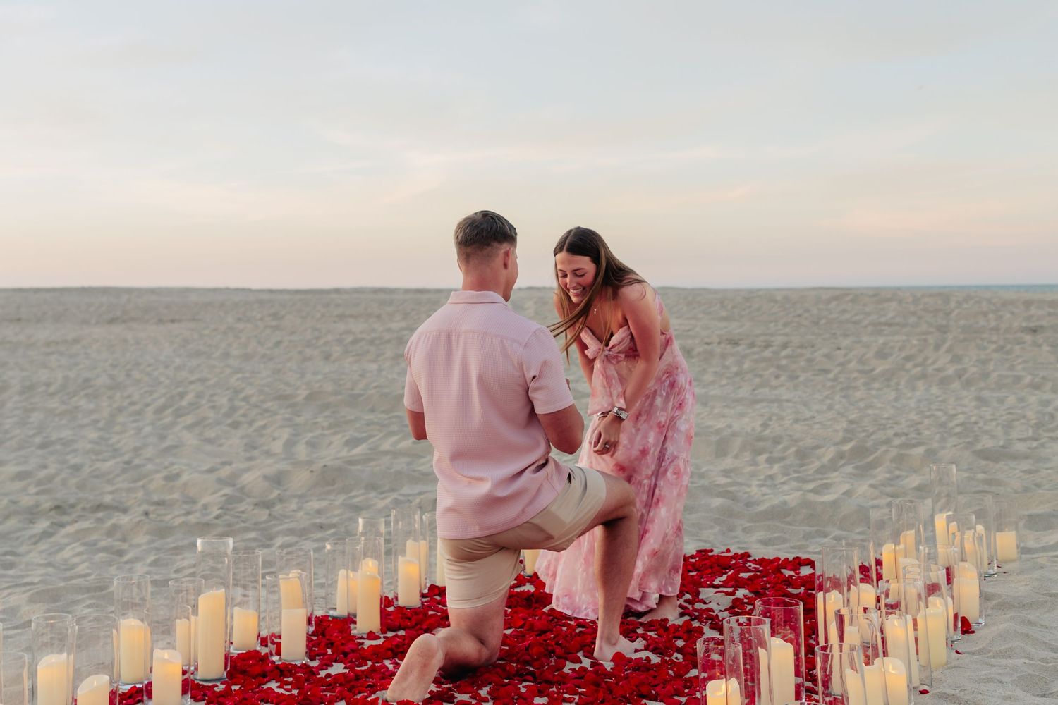 A romantic beach proposal scene with candles and rose petals arranged in a pathway at sunset.