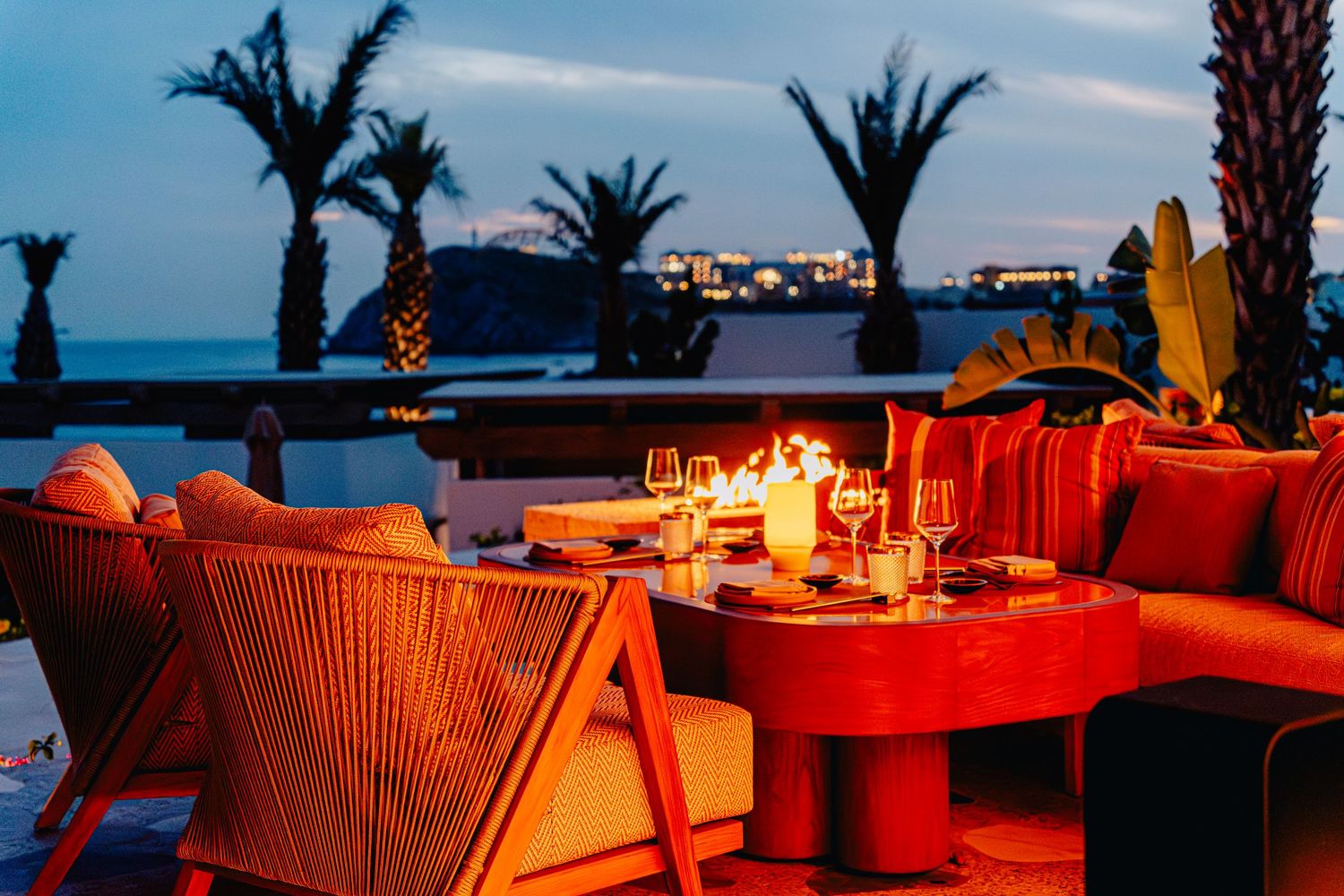 Romantic beachfront dining setup with candlelit tables and orange chairs at sunset.