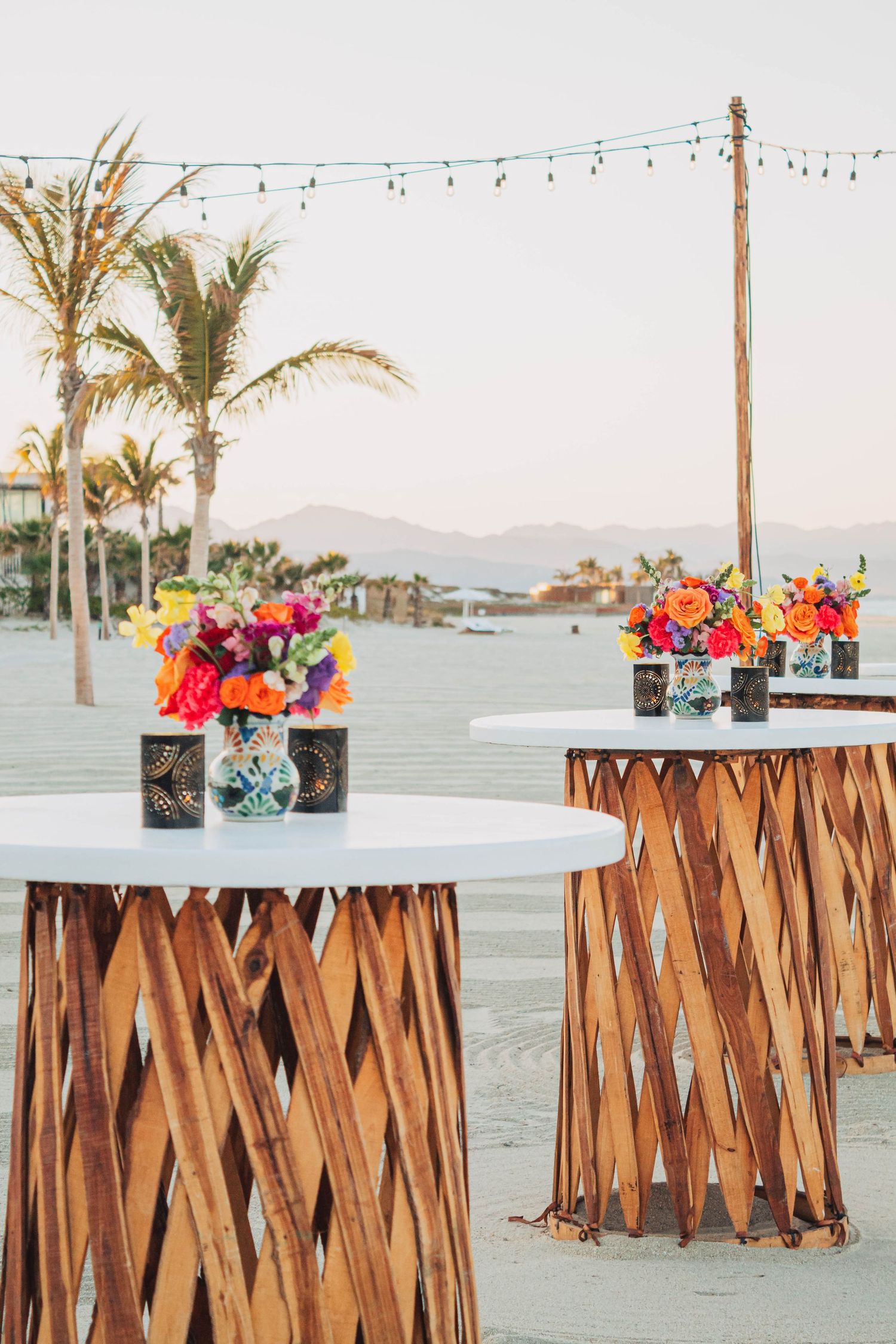 Stylish cocktail tables with woven bases and vibrant floral arrangements under string lights.