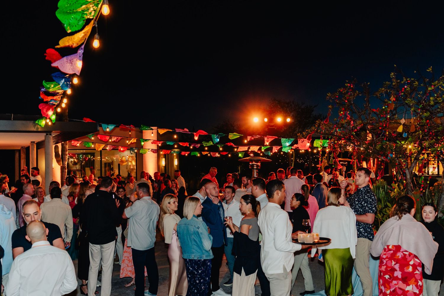 A festive outdoor party at night with colorful string lights and a crowd of people dancing and socializing.