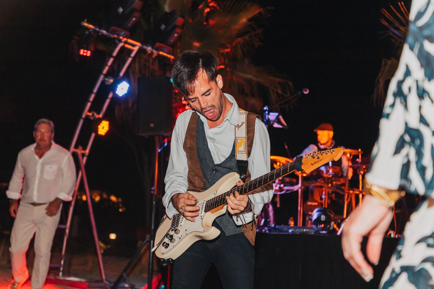 A performer plays electric guitar on stage during a concert with colorful stage lighting and band members in the background.