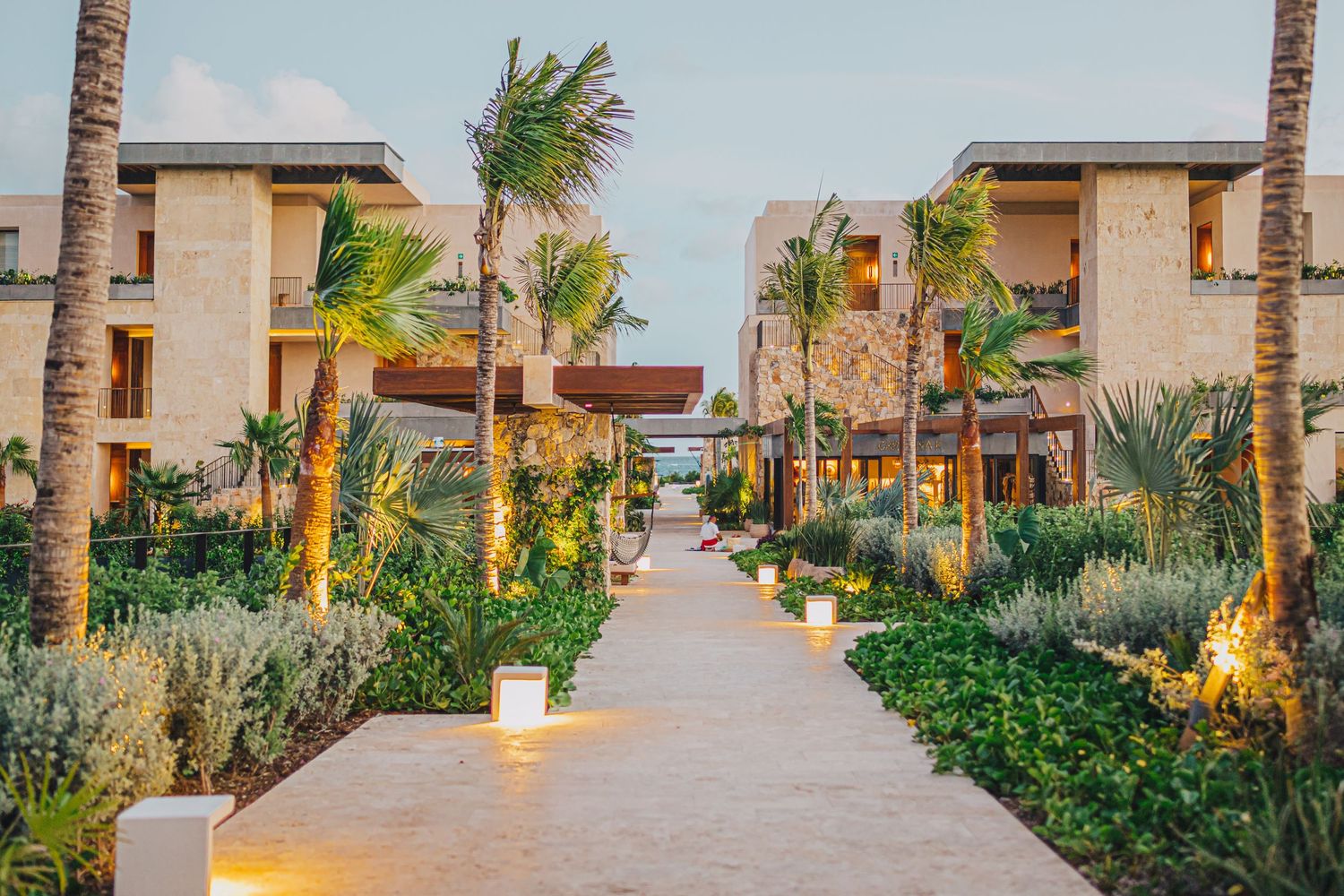 Evening view of a palm-lined pathway leading through a luxury resort complex with illuminated buildings and landscaping.