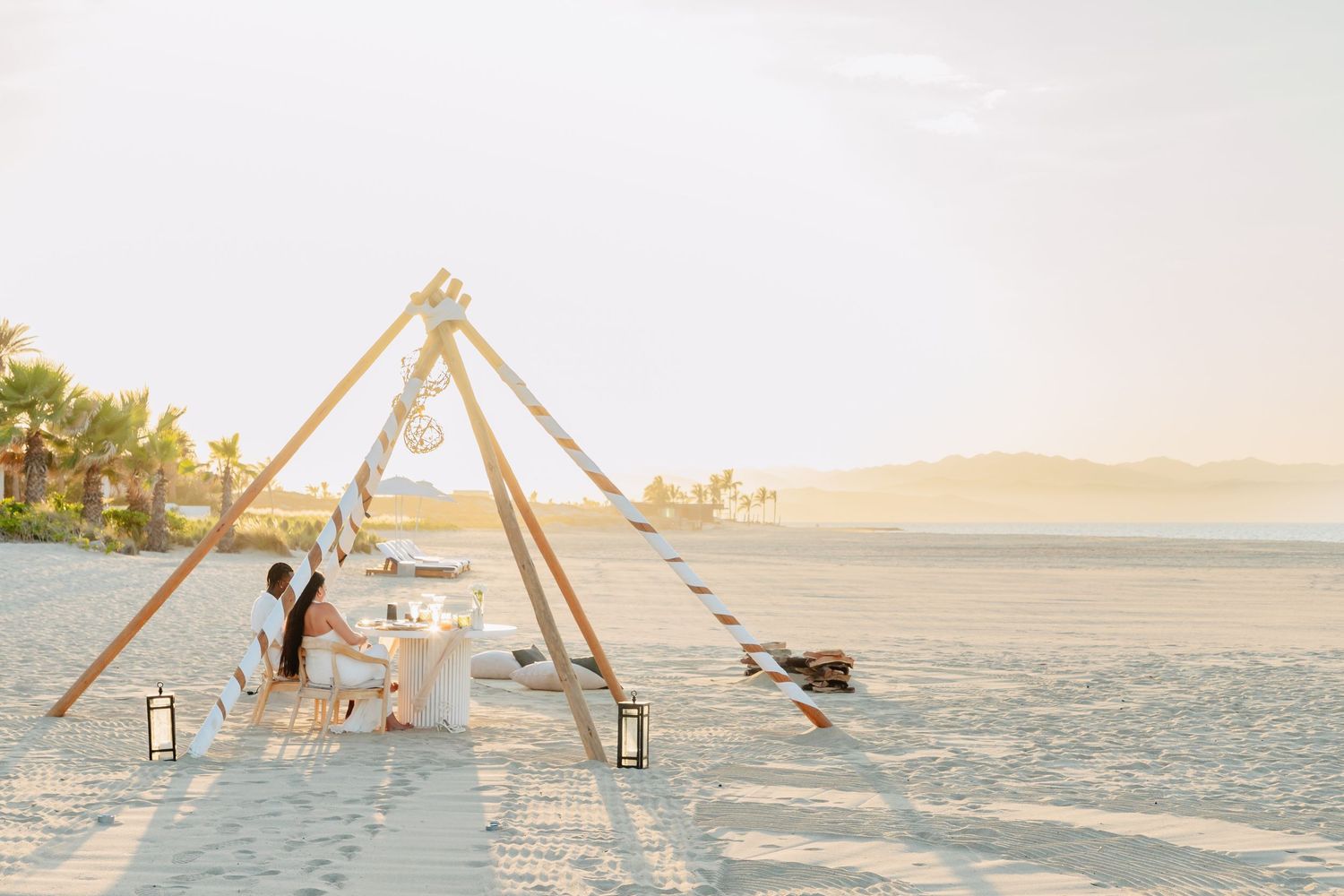 A wooden A-frame structure stands on a pristine white sandy beach at sunset with ocean views.