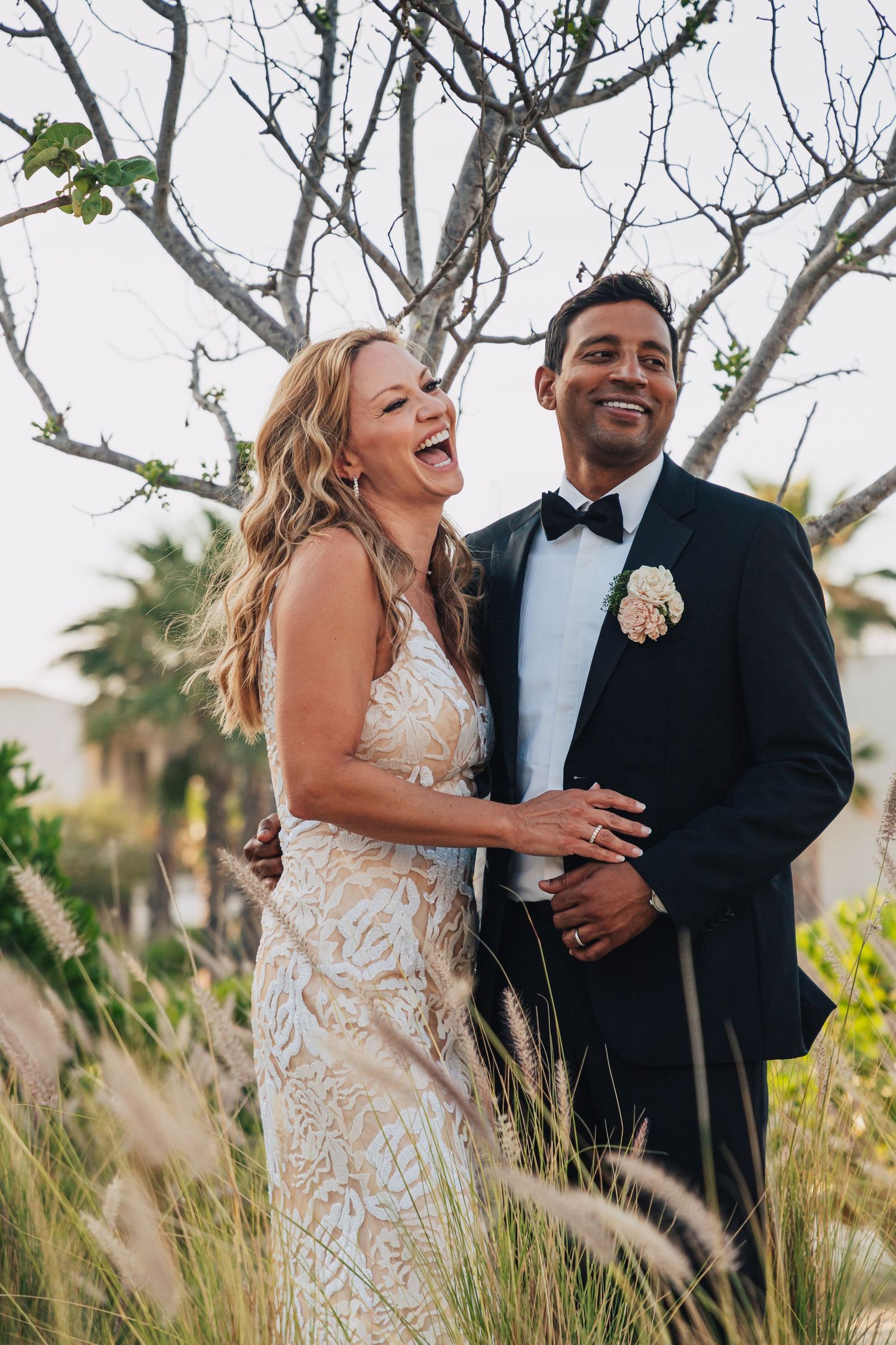 A joyful bride and groom share candid laughter together in an outdoor setting with tall grass and trees.